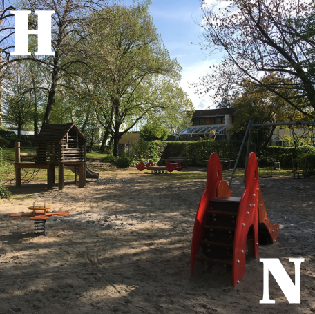 Foto des aktuellen Kinderspielplatzes. In einem großen Sandbereich stehen ein Spielhaus, eine Minirutsche und eine Miniwippe. Weiter hinten auf dem Gelände steht eine Schaukel, das Gebiet ist umgeben von Bäumen und Büschen.