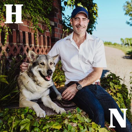 Ein Mann sitzt auf einer Mauer und lächelt in die Kamera. Neben ihm liegt ein Hund. Der Mann hat seinen rechten Arm um den Hund gelegt. Im Hintergrund sind eine pflanzenbewachsene Backsteinmauer und Weinstöcke zu sehen. 