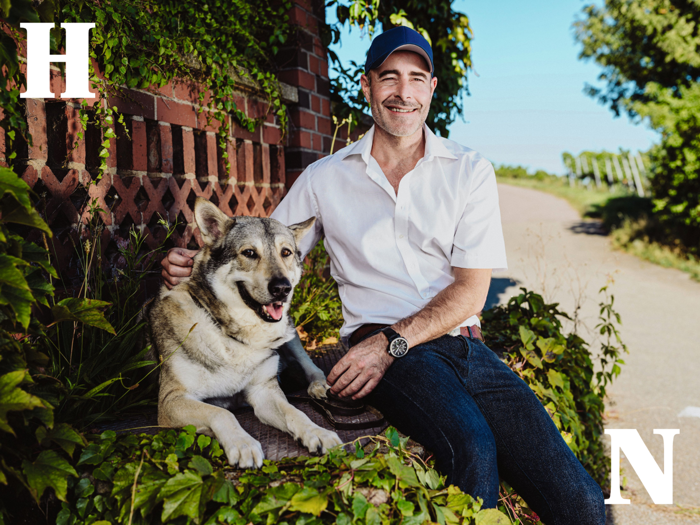 Ein Mann sitzt auf einer Mauer und lächelt in die Kamera. Neben ihm liegt ein Hund. Der Mann hat seinen rechten Arm um den Hund gelegt. Im Hintergrund sind eine pflanzenbewachsene Backsteinmauer und Weinstöcke zu sehen. 