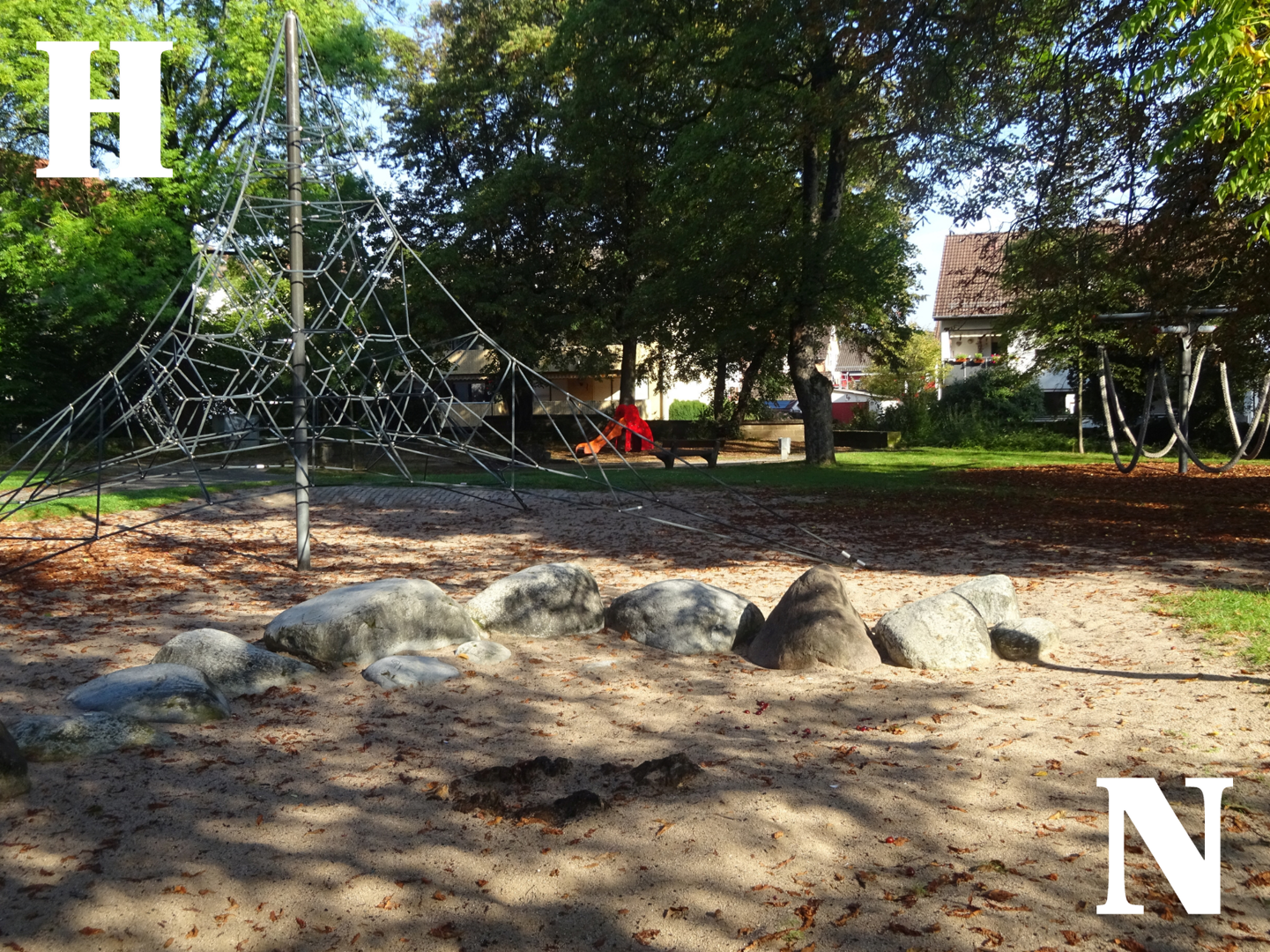 Foto des aktuellen Kinderspielplatzes. Im Vordergrund liegen große Natursteine, die den Sandbereich abgrenzen. Dahinter steht ein großes Seilklettergerüst. Im Hintergrund stehen eine rote Kleinkindrutsche und eine Parkbank. Sie sind umgeben von Rasenfläche und hochgewachsenen Bäumen.