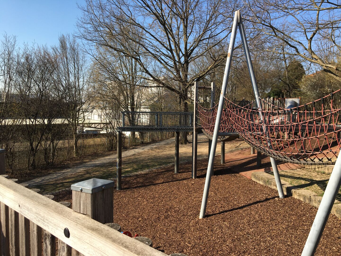 Zu sehen ist der aktuelle Spielplatz Kohlpfad in der Gruppenbacher Straße. Im Vordergrund steht eine Hängebrücke, im Hintergrund grenzt eine Plattform auf Stelzen an. Rundherum ist der Spielplatz mit zahlreichen hochgewachsenen Bäumen gesäumt.