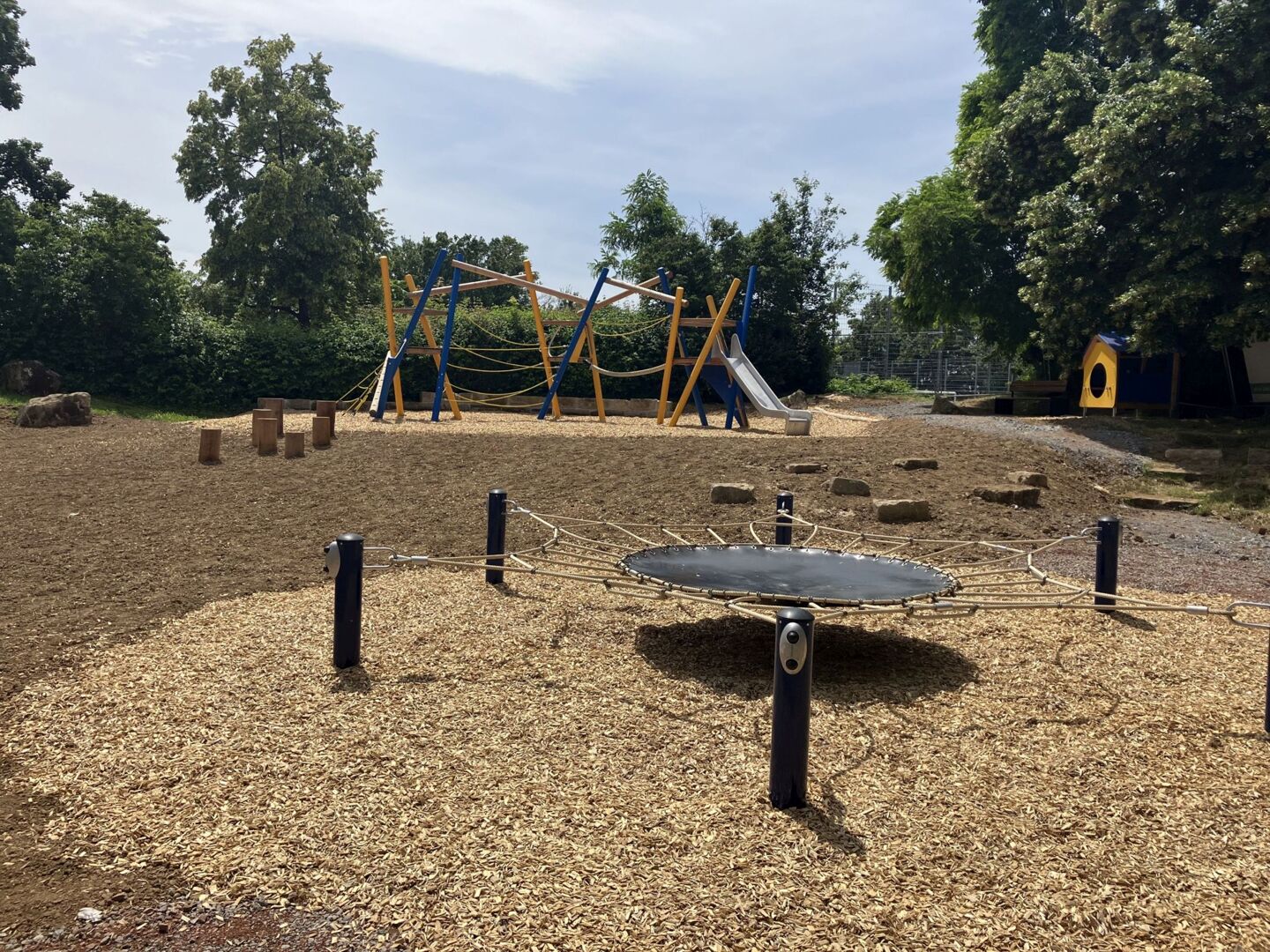 Foto des sanierten Spielplatzes. Im Vordergrund ist ein Sprungnetz zu sehen, im Hintergrund ein großes Klettergerüst mit Seilen, einem Kletternetz und einer Rutsche sowie ein Spielhäuschen für kleinere Kinder. 
