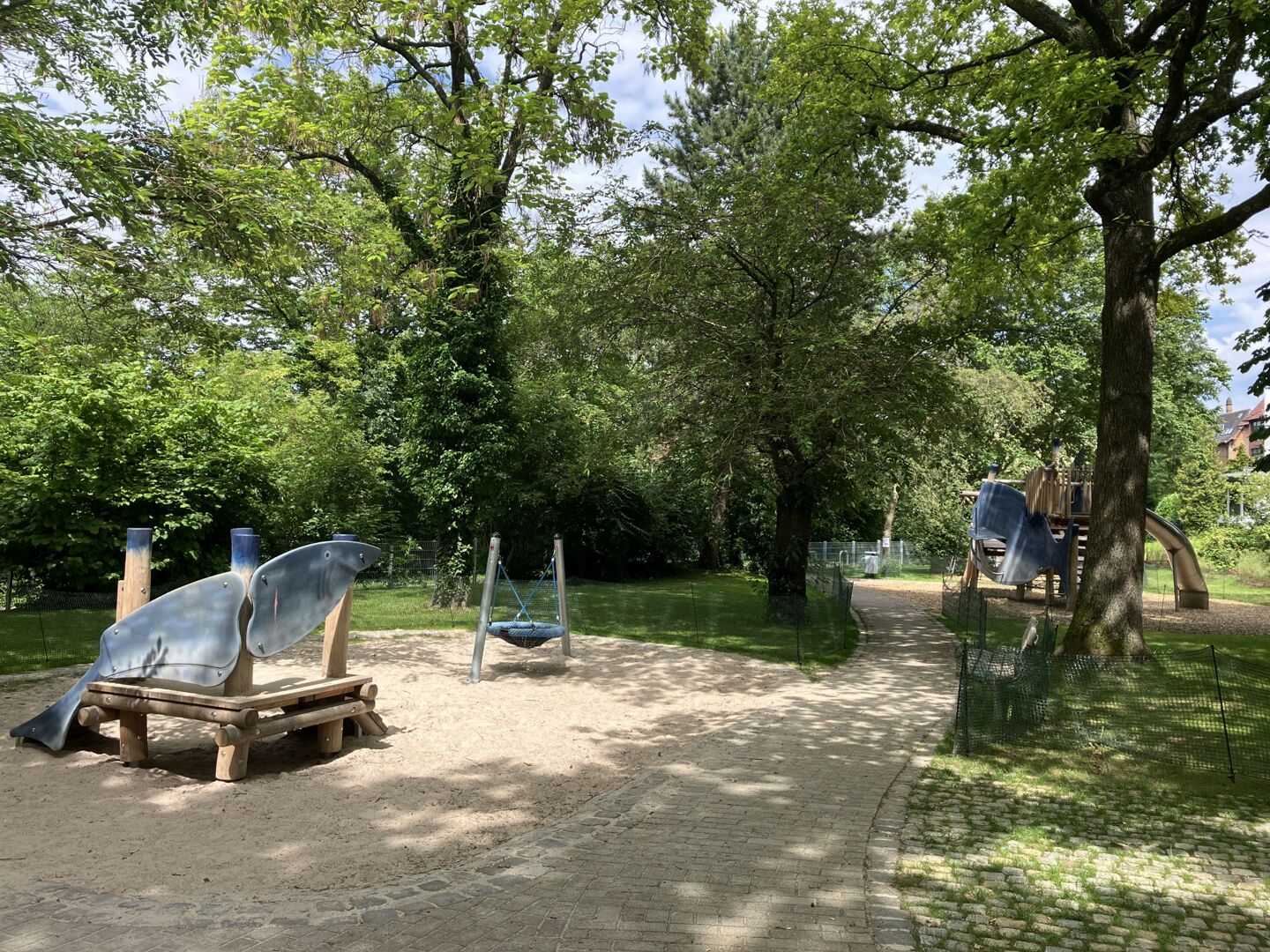 Foto des aktuellen Spielplatzes. Im Vordergrund befindet sich eine Mini-Rutsche für Kleinkinder in der Optik eines Haifischs. Rechts daneben eine Mini-Nestschaukel. Beide Spielgeräte stehen in einem Sandkasten. Ein gepflasterter Weg führt in den hinteren Bereich des Spielplaztes zu einem großen Klettergerüst mit Rutsche, ebenfalls in Haifischoptik. Rundherum stehen viele hochgewachsene Bäume auf Rasenflächen.