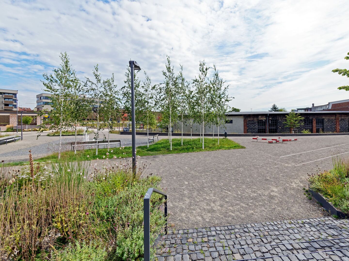 Foto des fertiggestellten ersten Bauabschnitts des Bahnbogens Süd. Eine Treppe aus Pflastersteinen führt auf den Platz. Auf einer schmalen Rasenfläche stehen Birken, dahinter sind auf einem gepflasteren Platz mit Wasserspiel Sitzbänke aufgestellt. Im Hintergrund sind die neue Böckinger Mitte, die überdachte S-Bahn-Haltstelle des Sonnenbrunnens und die Rückseite des Ringlokschuppens zu sehen.